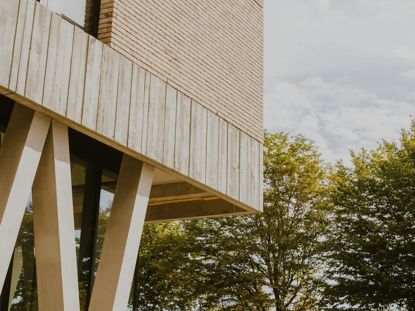 Corner of a modern brick building resting on angled concrete columns, with trees beside it